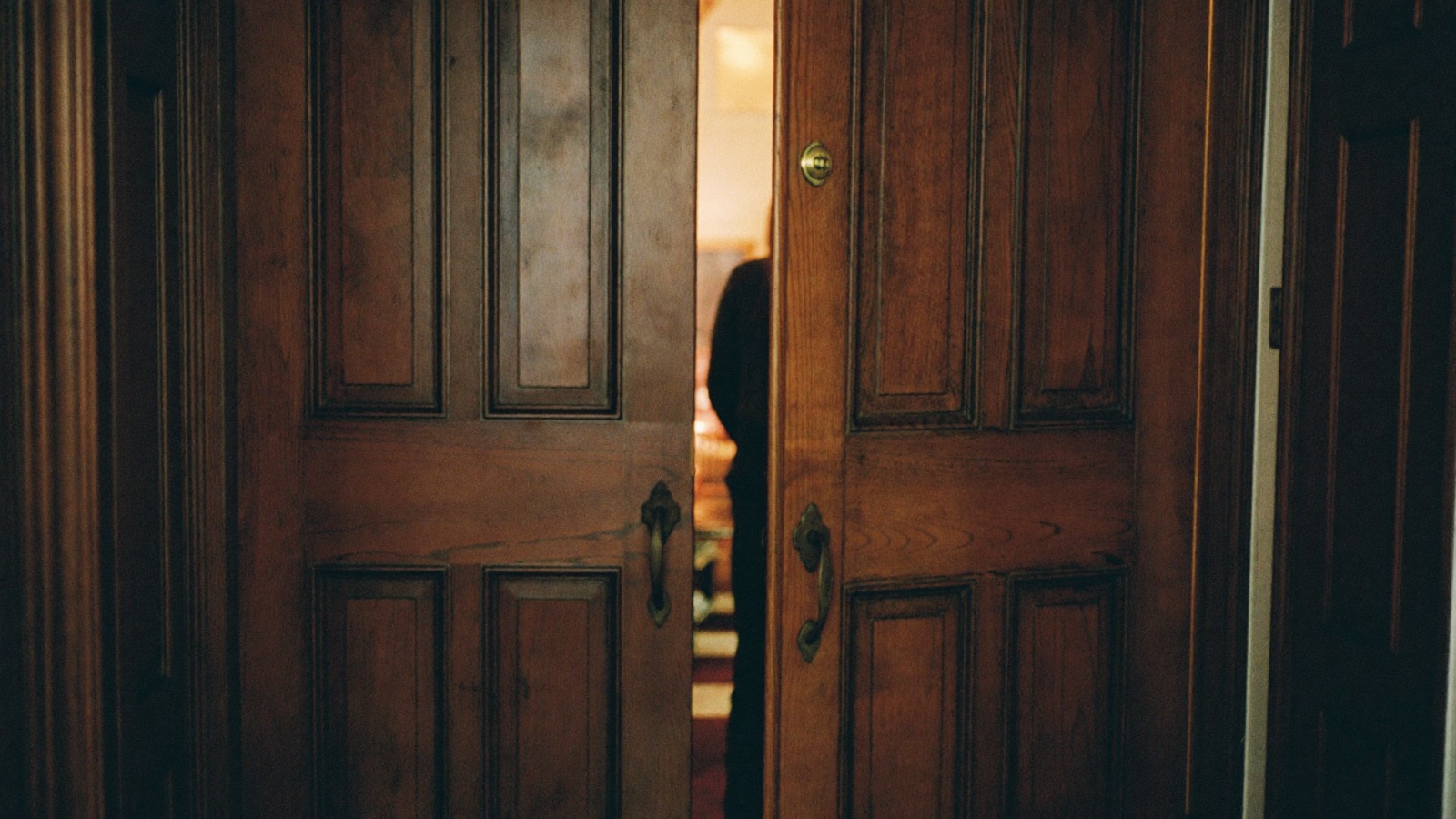 A heavy door ajar in a dim hallway, the light of a lively room spilling through.