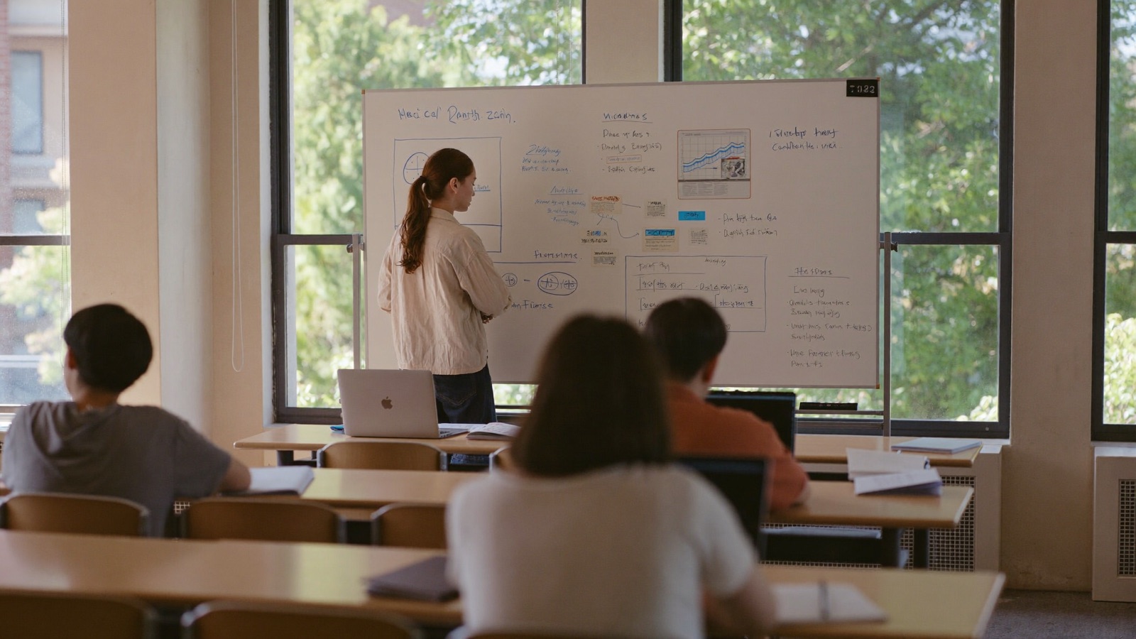 A small seminar room with a whiteboard full of diagrams and a few students at laptops.