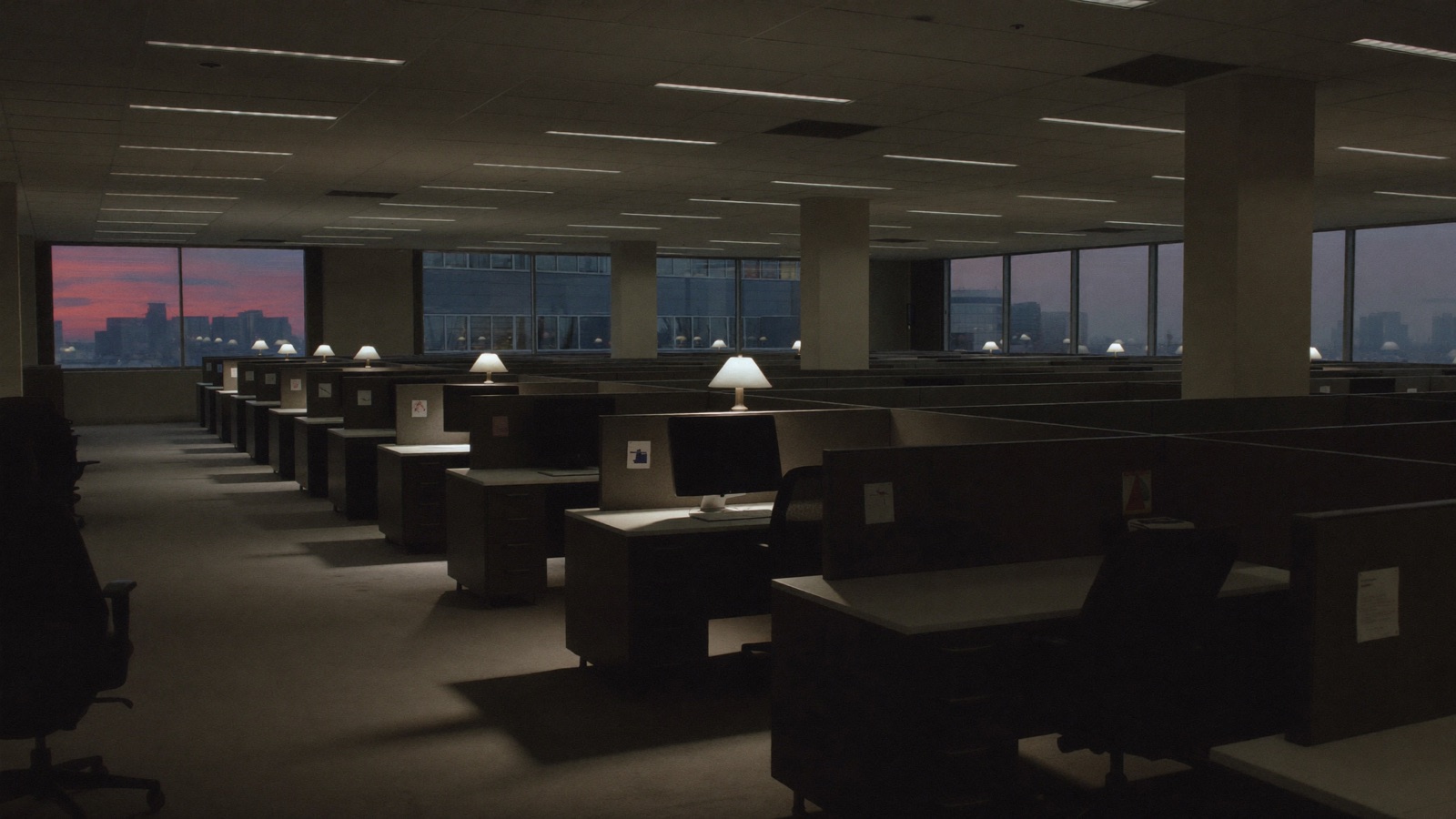 An empty office floor at dusk, rows of unused desks and one lamp on.